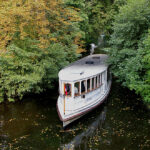 Die weiße Alsterbarkasse 'St. Georg' fährt auf einem schmalen Kanal, der von dichtem grünem Laub umgeben ist.