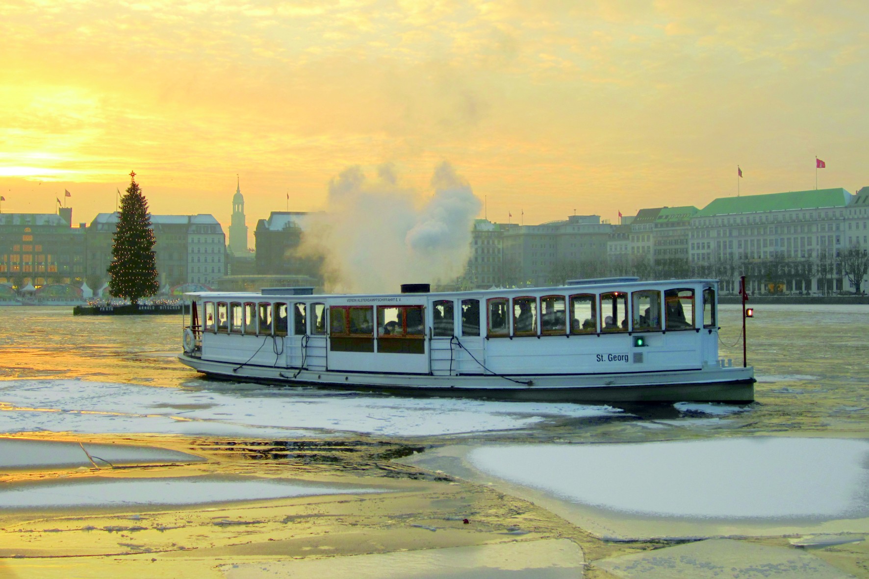 Die ST. GEORG fährt bei winterlichem Abendlicht durch die teils vereiste Alster, im Hintergrund die beleuchtete Alstertanne.