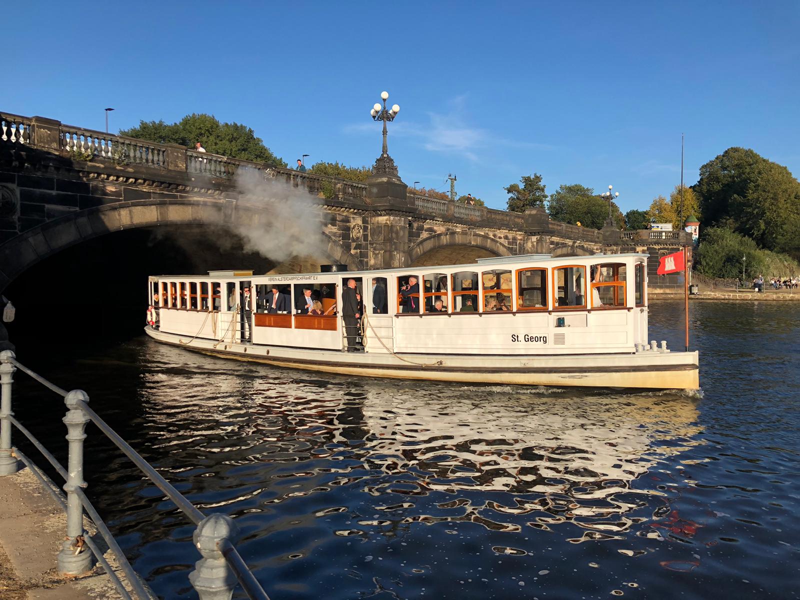 Die Alsterbarkasse 'St. Georg' fährt unter einer steinernen Brücke hindurch, mit Passagieren an Bord und dampfenden Schornstein.