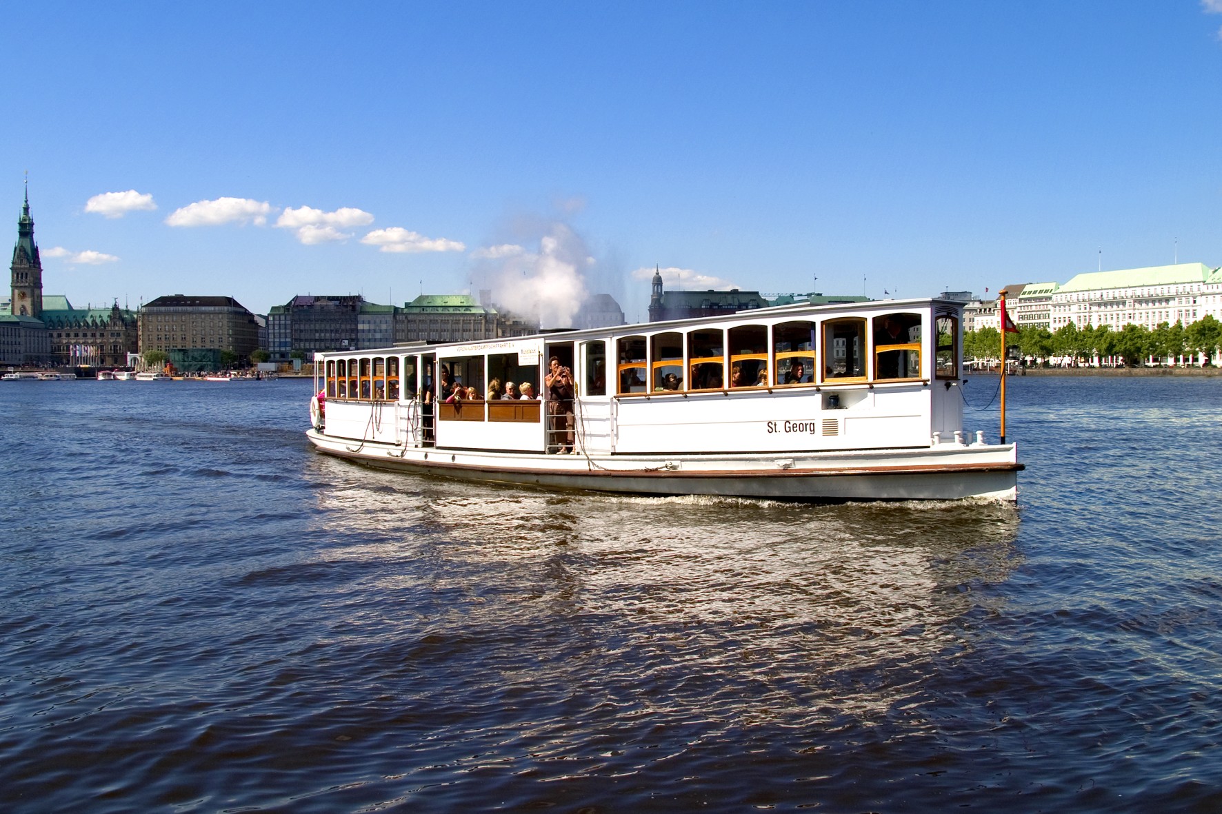 Ein weißes Alsterdampfschiff, die 'St. Georg', fährt bei sonnigem Wetter über die Alster, im Hintergrund ist die Uferpromenade von Hamburg zu sehen.