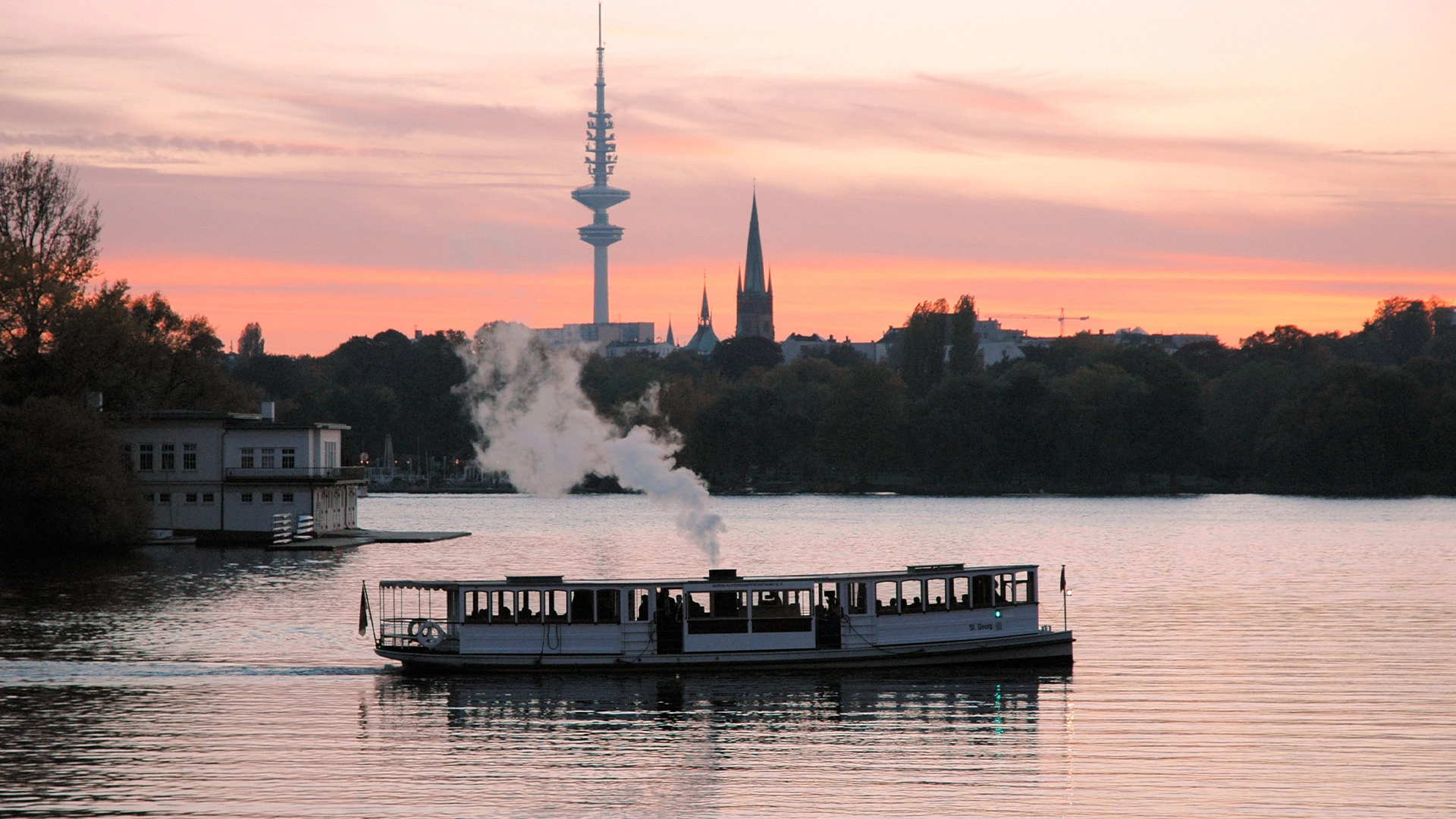 Eine Barkasse fährt auf der Außenalster in Hamburg bei Sonnenuntergang, mit dem Fernsehturm im Hintergrund.