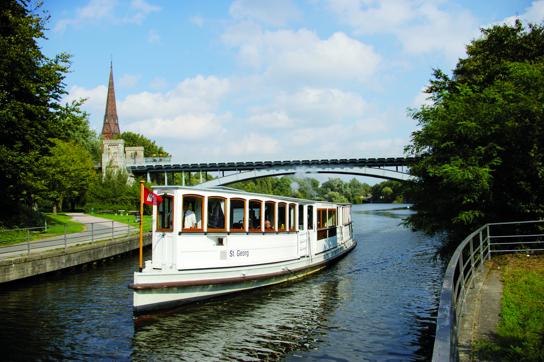 Das Alsterdampfschiff 'St. Georg' fährt unter einer Brücke hindurch, umgeben von grünen Bäumen an den Ufern.