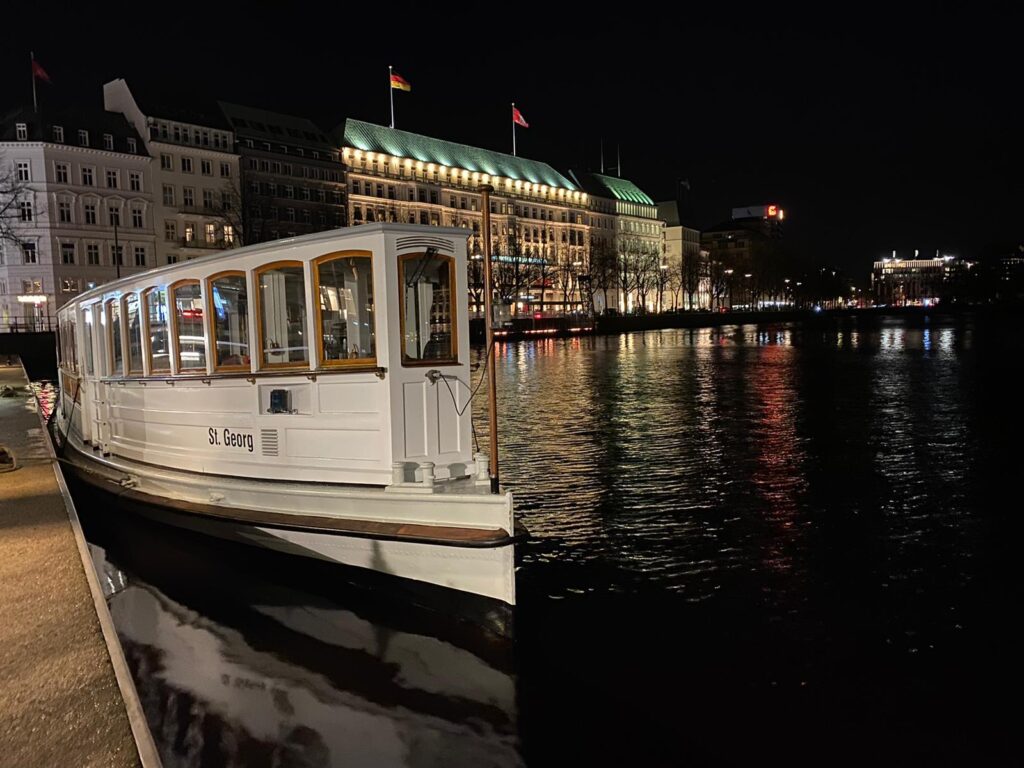 Die Alsterbarkasse 'St. Georg' liegt nachts beleuchtet am Ufer, im Hintergrund leuchtet das Hotel Vier Jahreszeiten.