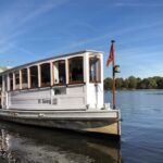 Ein weißes Alsterdampfschiff, die 'St. Georg', fährt bei klarem Wetter über das Wasser, im Hintergrund ist die Uferlandschaft zu sehen.