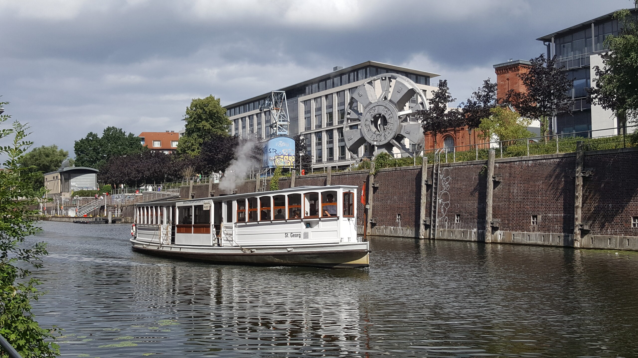 Die Alsterbarkasse 'St. Georg' fährt auf einem Kanal in Hamburg an modernen Gebäuden und alten Industriebauten vorbei.