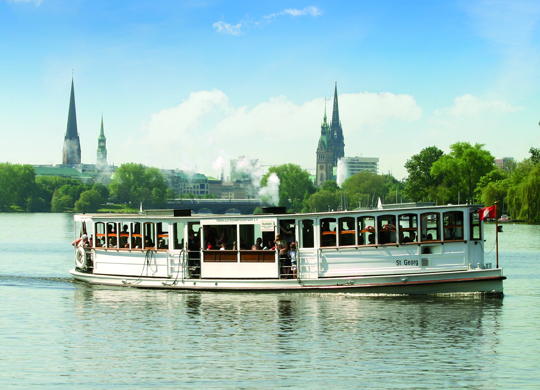 Ein weißes Alsterdampfschiff, die 'St. Georg', fährt bei sonnigem Wetter über das Wasser, im Hintergrund ist die Hamburger Skyline mit Kirchtürmen und dem Rathaus erkennbar.
