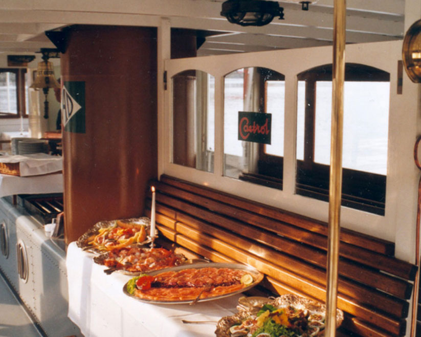 Ein gedecktes Buffet mit verschiedenen Speisen steht an der Seite eines historischen Alsterdampfschiffs mit Holzbänken und Fenstern. Ein Schild mit der Aufschrift 'Castrol' ist an einer Tür zu sehen.