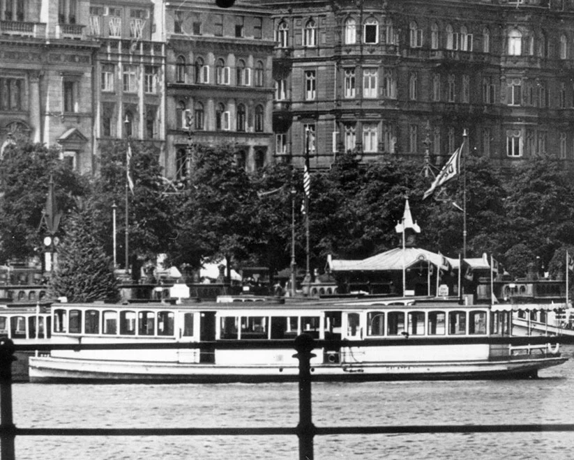 Ein historisches Schwarz-Weiß-Foto eines großen Alsterdampfers mit Passagieren, der auf dem Wasser fährt. Im Hintergrund sind historische Gebäude und Bäume zu sehen.