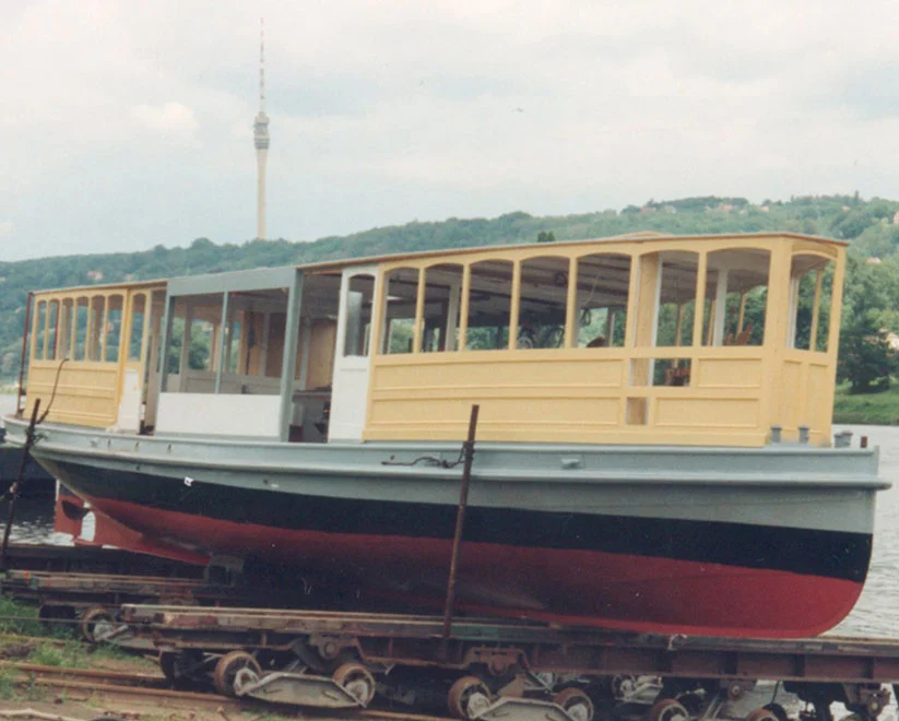Eine historische Barkasse mit gelbem Aufbau steht auf einem Rollwagen an Land, der rote Schiffsrumpf ist deutlich zu sehen. Im Hintergrund ragt der Hamburger Fernsehturm über die Baumgrenze.