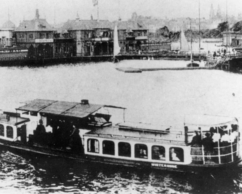 Ein historisches Schwarz-Weiß-Foto, das ein Alsterdampfschiff namens 'Winterhude' auf dem Wasser zeigt, mit Passagieren an Bord. Im Hintergrund ist eine Anlegestelle mit Gebäuden zu sehen.