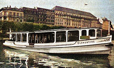 Eine historische, farbige Aufnahme des Alsterdampfschiffs 'Clementine', das mit Passagieren an Bord vor der bebauten Uferpromenade von Hamburg fährt.