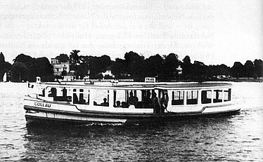 Ein historisches Schwarz-Weiß-Foto zeigt ein Alsterdampfschiff namens 'Kollau' mit Passagieren an Bord, das bei trübem Wetter über das Wasser fährt.